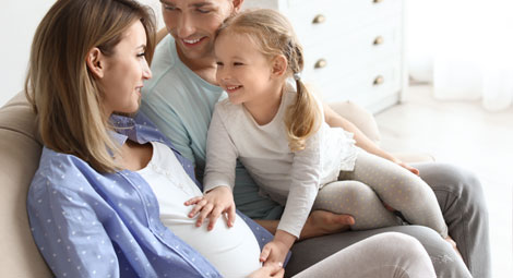 A family sat on a sofa together. The mother is pregnant and a little girl is touching her stomach