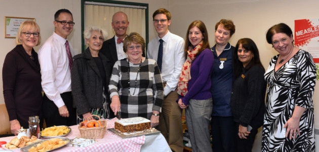 Group of people around a table with food. A lady is cutting a cake